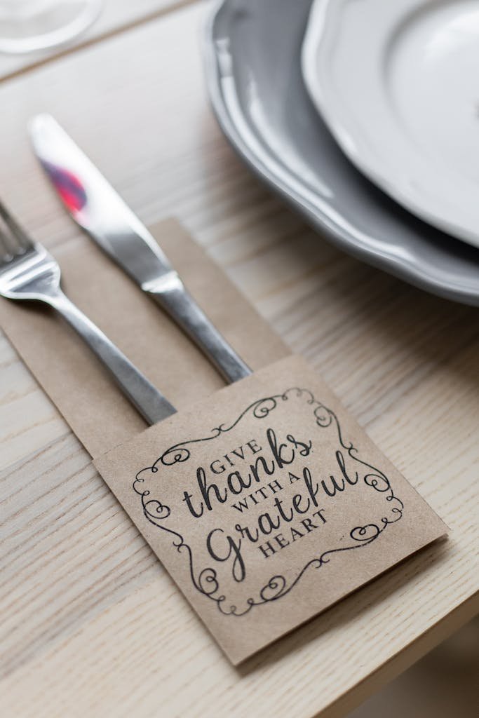 From above of metal shiny fork and knife placed in paper cover with inscription for Thanksgiving near plates on wooden table