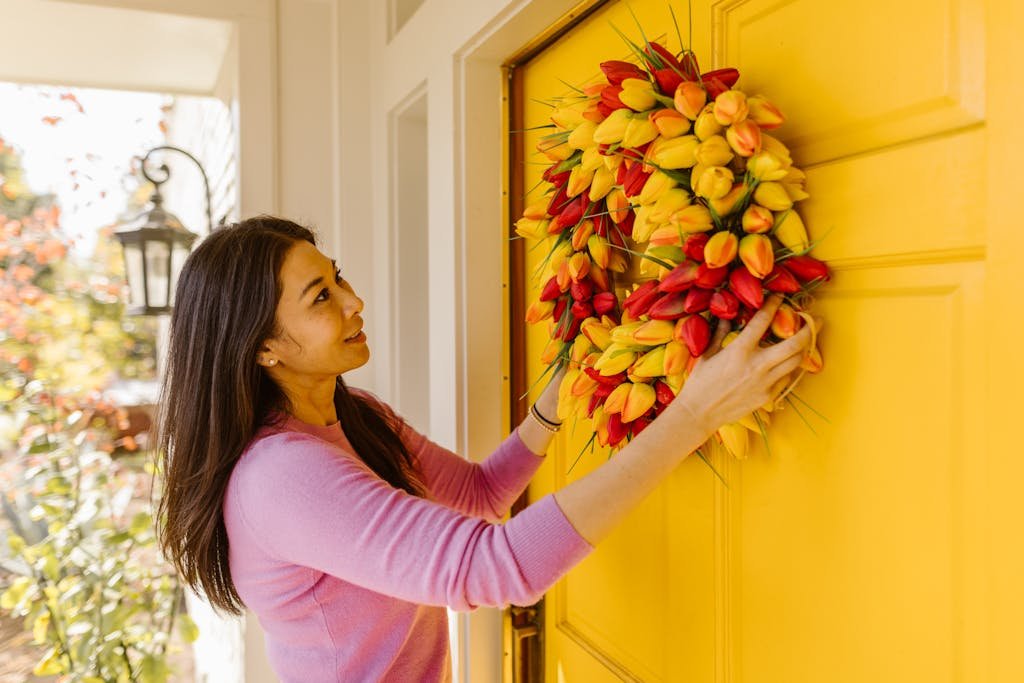 A woman hangs a vibrant floral wreath on a bright yellow door, symbolizing spring decor.