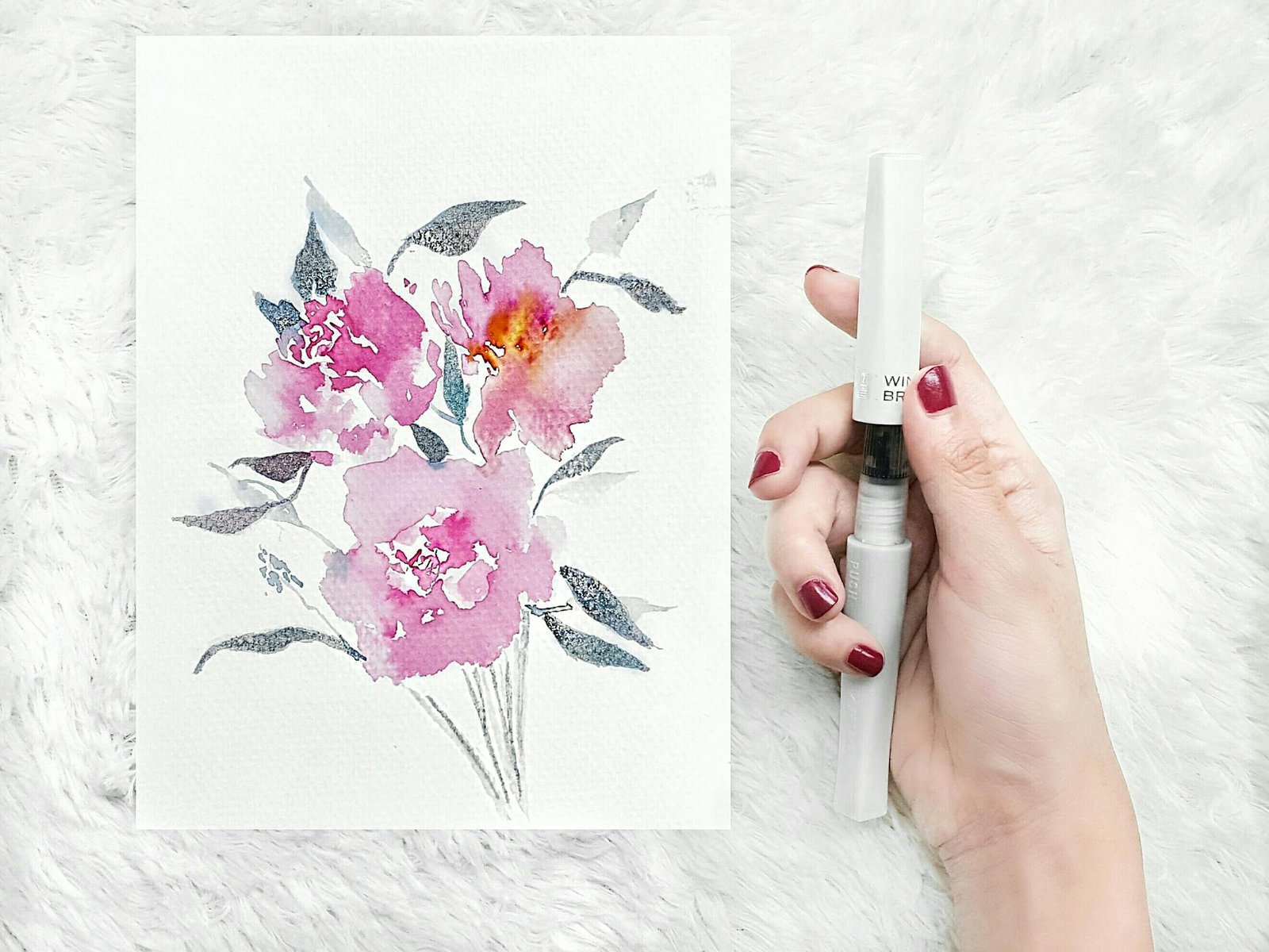 A hand holding a watercolor brush next to a floral painting on paper.